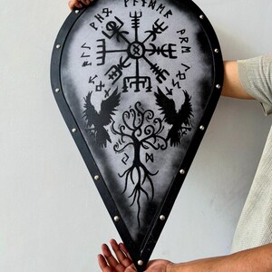 May include: A teardrop-shaped shield with a black border and silver-toned rivets. The center features a white background with a black Vegvisir symbol, two ravens, and a tree of life design. The shield is decorated with black runic inscriptions.