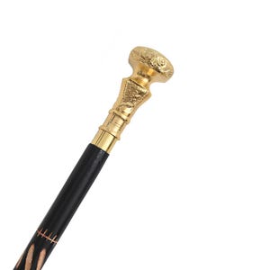 May include: A walking stick featuring a polished black shaft with carved details and a gold-colored metal handle. The handle has an ornate, embossed design with a rounded top and a tapered neck that connects to the dark, smooth wooden shaft.
