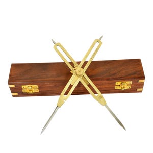 May include: A brass proportional divider tool with sharp pointed tips and a central adjustment screw, positioned in front of a rectangular dark brown wooden storage box featuring two gold-colored metal latches and brass corner accents.
