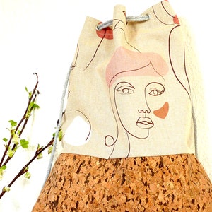 May include: A beige drawstring bag with a cork bottom. The bag has a graphic design of a woman's face with pink and orange accents.