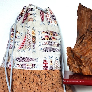 Puede incluir: Una bolsa con cordón con base de corcho y parte superior de tela beige. La tela está estampada con diseños de peces coloridos en rojo, azul y amarillo. El cordón es gris. La bolsa se muestra sobre un fondo blanco.