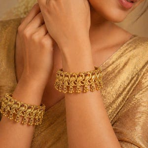 May include: Gold-toned bangles with intricate peacock and floral designs. The bangles feature small red accents and dangling embellishments. The jewelry is displayed on a person's arm, showcasing the ornate details.
