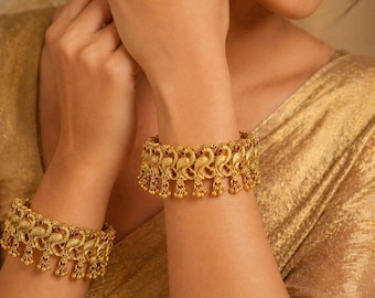 Puede incluir: Brazaletes dorados con intrincados diseños de pavo real y flores. Los brazaletes presentan pequeños detalles rojos y adornos colgantes. Las joyas se muestran en un brazo, mostrando los detalles ornamentados.