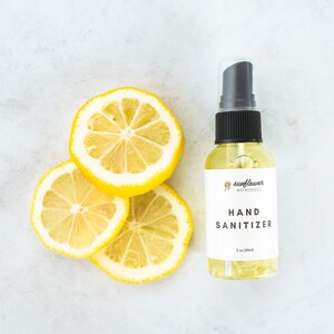 Puede incluir: Una botella de pl&aacute;stico transparente de desinfectante de manos con una boquilla de pulverizaci&oacute;n negra y una etiqueta blanca que dice "sunflower motherhood hand sanitizer 2 oz (60ml)". Tres rodajas de lim&oacute;n est&aacute;n dispuestas sobre una superficie de m&aacute;rmol blanco.