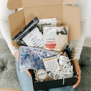 May include: A black box filled with various items, including a blue ice pack, a black and white card with the text "Life is messy but you don't have to be.", a clear spray bottle labeled "Mama Oil", a small tin labeled "sunflower", a black and white card with the text "Mama Affirmations", a black and white card with the text "UpSpring CPANTY post c-section", and a black and white card with the text "sunflower".