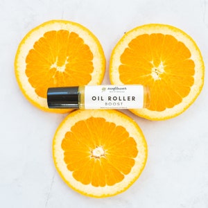 Puede incluir: Un frasco de rodillo blanco con tapa negra y una etiqueta que dice "Sunflower Botanicals Oil Roller Boost" se coloca sobre tres rodajas de naranja.
