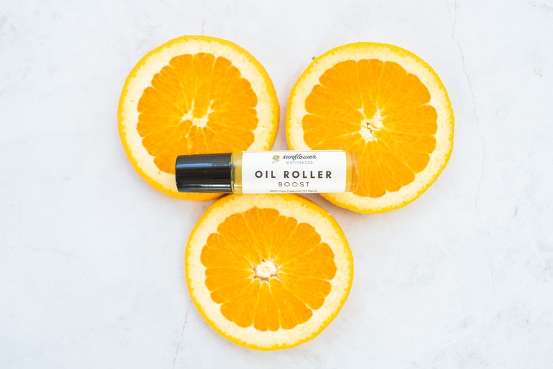 Puede incluir: Un frasco de rodillo blanco con tapa negra y una etiqueta que dice "Sunflower Botanicals Oil Roller Boost" se coloca sobre tres rodajas de naranja.
