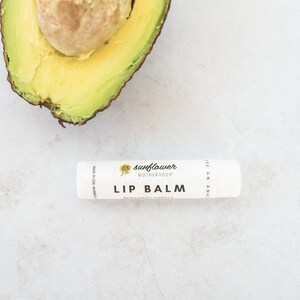 Puede incluir: Un tubo de b&aacute;lsamo labial blanco con el texto "sunflower motherhood" y "lip balm" impreso en la etiqueta. El b&aacute;lsamo labial est&aacute; sobre una superficie blanca con la mitad de un aguacate en el fondo.
