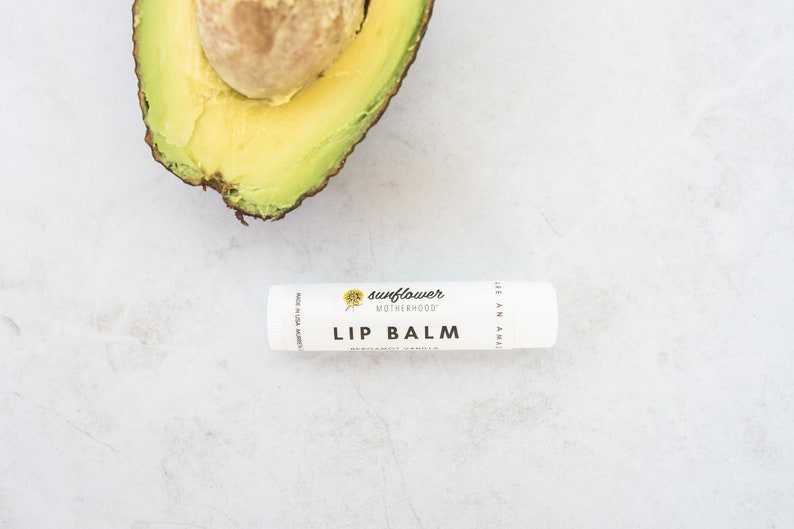 Puede incluir: Un tubo de b&aacute;lsamo labial blanco con el texto "sunflower motherhood" y "lip balm" impreso en la etiqueta. El b&aacute;lsamo labial est&aacute; sobre una superficie blanca con la mitad de un aguacate en el fondo.