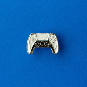 Op de afbeelding: Een wit en zwarte videogamecontroller-pin met gouden rand, tegen een levendige blauwe achtergrond. De pin heeft gedetailleerde knop- en joystickontwerpen, die de essentie van moderne gaming-accessoires vastleggen.