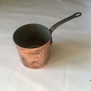 Puede incluir: Una cacerola de cobre con un mango largo de metal oscuro. La olla tiene forma cilíndrica con un borde ligeramente ensanchado. El interior de la olla tiene un tono verdoso y el exterior muestra signos de edad y uso.