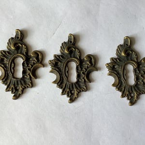 May include: Three antique brass keyhole escutcheons featuring an ornate, symmetrical leaf-like design with a central vertical opening. Each metal plate has a dark, weathered finish and small mounting holes at the top and bottom for attachment.