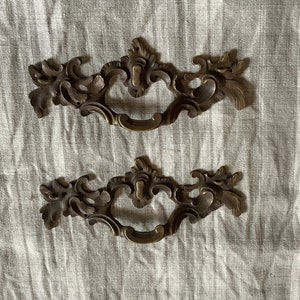 Può includere: Due tiretti per cassetti in ottone decorati con un design a rotolo. I tiretti hanno un'apertura centrale per un pomo o una maniglia.