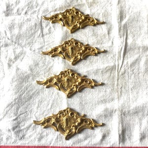 Peut inclure: Quatre plaques de serrure dorées ornées de volutes et d'un trou de serrure central. Disposées en rang sur une surface blanche texturée, elles mettent en valeur leur design élaboré et leur style vintage. Mesurent environ 10 cm de long.