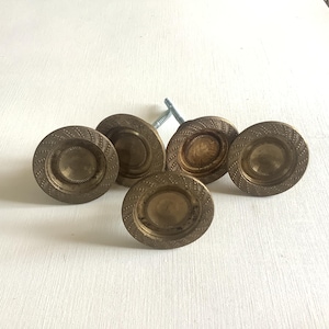 May include: Six antique brass cabinet knobs with a circular design and textured edges. Each knob has a central, slightly raised circle. The knobs are arranged on a white surface, with a single screw visible.