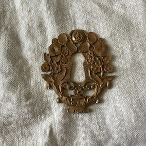 Pode incluir: Cobertura decorativa de buraco de fechadura em latão de estilo antigo. A cobertura tem a forma de uma coroa floral com um buraco de fechadura no centro. A coroa apresenta folhas, flores e uma rosa detalhadas. O latão tem uma pátina quente e envelhecida.