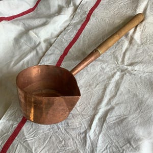 May include: A copper ladle with a wooden handle. The ladle has a rounded bowl and a spout for pouring. The handle is attached to the bowl with a copper fitting. The ladle is resting on a white cloth with red stripes.