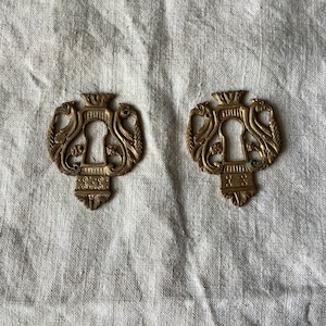 May include: Two antique brass keyhole escutcheons with ornate designs featuring a crown and two birds on each side.