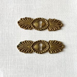 Puede incluir: Dos bocallaves antiguos de latón. Cada bocallave tiene un ojo de cerradura ovalado en el centro, rodeado por un borde en forma de cuerda. Los extremos de los bocallaves están adornados con un diseño de hoja estilizado. El latón tiene una pátina cálida y envejecida.