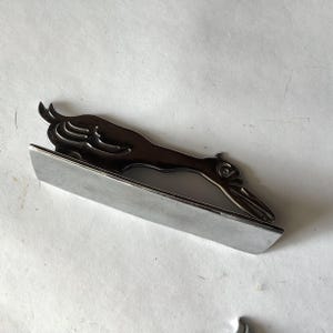 Pode incluir: Um prendedor de gravata prateado com um design de p&aacute;ssaro estilizado. O p&aacute;ssaro tem um bico longo e est&aacute; pousado sobre uma base retangular. O prendedor &eacute; feito de metal e tem um acabamento brilhante, adequado para ocasi&otilde;es formais. Mede cerca de 7 cm de comprimento.