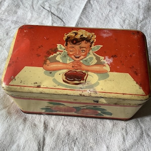 Peut inclure: Boîte en étain vintage avec un design rouge et jaune. Un jeune garçon est représenté souriant et regardant une assiette de pain avec de la confiture. La boîte a un aspect vintage usé.