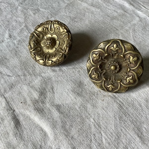 Könnte beinhalten: Zwei antike Schubladengriffe aus Messing mit floralen Designs. Die Griffe sind rund mit detaillierten, erhabenen Mustern aus Blumen und Blättern. Das Messing hat eine warme, gealterte Patina, die auf Vintage- oder antike Ursprünge hindeutet.