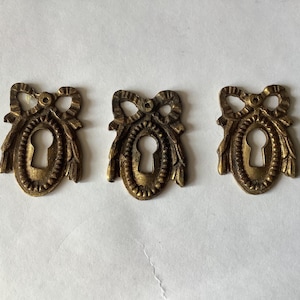 Op de afbeelding: Drie antieke messing sleutelgatdeksels. Elke afdekking heeft een ovale vorm met een sleutelgat in het midden, omgeven door een decoratieve rand. Bovenop elke afdekking bevindt zich een strik. Ze zijn waarschijnlijk voor meubels of deuren.