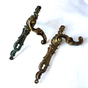 May include: Two ornate brass door handles with intricate scrollwork and keyhole plates. One handle features a bright metallic gold finish, while the other displays a weathered, dark green patina. Both pieces have curved levers and mounting holes.