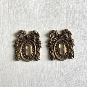 Könnte beinhalten: Zwei antike Schlüssellochabdeckungen aus Messing. Jede Abdeckung hat eine ovale Form mit einem Schlüsselloch in der Mitte, umgeben von einem dekorativen Rand und gekrönt von einer Schleife und floralen Akzenten. Das Messing hat eine warme, gealterte Patina.