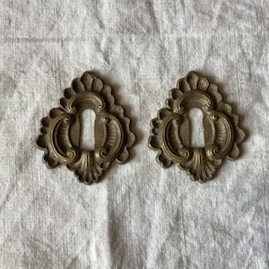 Può includere: Due copri-chiavi in ​​ottone decorati con elaborate volute e un buco della serratura al centro.