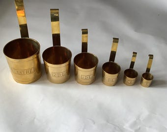 May include: Set of six brass measuring cups with long handles. The cups are labeled with measurements such as "LITRE", "2DL", "1DL", "DL", "CL", and "CL". The cups are arranged in descending order by size, with the largest cup on the left.