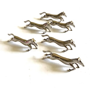 Könnte beinhalten: Ein Set aus sechs silberfarbenen Metallpferdefiguren, die jeweils in einer dynamischen Galopppose mit ausgestreckten Beinen und gewölbtem Rücken dargestellt sind. Die Pferde haben glatte, reflektierende Oberflächen und detaillierte Mähnen.