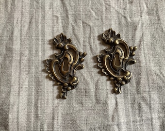 May include: Two ornate, gold-toned metal keyhole covers with intricate floral designs. The keyhole covers are on a light gray fabric background.