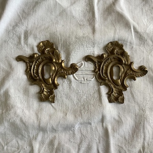 Op de afbeelding: Twee sierlijke, goudkleurige metalen sleutelgatbedekkingen met ingewikkelde ontwerpen. De sleutelgatbedekkingen bevinden zich op een witte stoffen achtergrond.