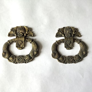 May include: Two matching brass drawer pulls featuring an ornate, circular drop handle design. Each piece displays intricate floral and leaf relief detailing on the mounting plate and the swinging ring, finished with a darkened, aged metallic patina.