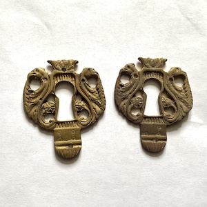 May include: Two antique brass keyhole covers. Each cover has an ornate design with a keyhole shape in the centre, flanked by detailed carvings of mythical creatures and topped with a crown. The brass has a vintage patina.