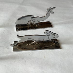 Pode incluir: Duas esculturas de metal cromado. Uma retrata um p&aacute;ssaro com o bico aberto, a outra um coelho correndo. Ambas s&atilde;o montadas em bases retangulares. As esculturas t&ecirc;m uma apar&ecirc;ncia met&aacute;lica reflexiva.