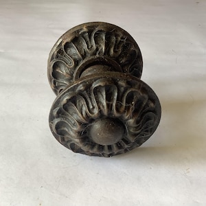 Puede incluir: Un par de pomos de puerta antiguos, de color marrón oscuro, con detalles ornamentados en forma de flor. Los pomos tienen un aspecto envejecido, lo que sugiere edad y uso. El diseño presenta un elemento central redondeado rodeado de patrones curvos estilizados.