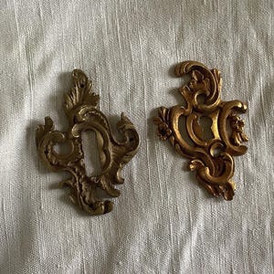 Op de afbeelding: Twee sierlijke, goudkleurige metalen sleutelgatbedekkingen met ingewikkelde krulontwerpen.
