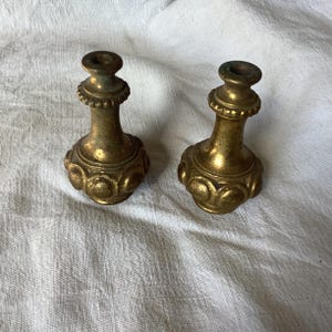 Op de afbeelding: Twee goudkleurige metalen eindstukken met een decoratief, versierd ontwerp. De eindstukken hebben een bolvormige vorm met een afgeronde bovenkant en een klein gaatje in het midden.