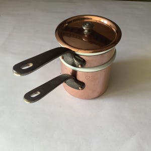 May include: Two copper saucepans with dark metal handles and a copper lid. The saucepans are stacked, with the smaller one on top. The rims are white. The lid has a small, round handle. The copper has a shiny, reflective surface.