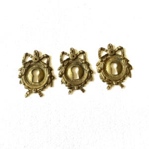 May include: Three antique brass keyhole covers. Each cover features a keyhole surrounded by a laurel wreath design, topped with a bow. The brass has a warm, aged patina, suggesting vintage or antique origins. These decorative hardware pieces are ideal for furniture restoration.