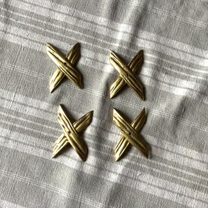May include: Four gold-colored, X-shaped decorative accents. Each accent has a textured surface and is arranged in a square formation against a gray and white striped background. The accents appear to be made of metal.
