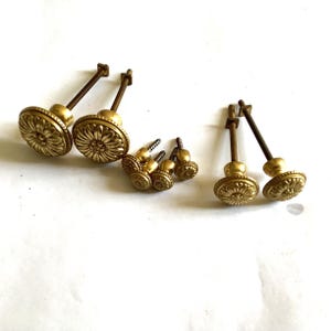 Puede incluir: Un conjunto de pomos de metal en tono latón con una cara circular que presenta un patrón floral o de rayos de sol en relieve. El set incluye varios pomos con vástagos metálicos roscados largos y algunos pomos más pequeños con tornillos cortos.