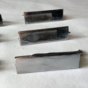 Pode incluir: V&aacute;rias puxadores de gaveta retangulares de metal prateado est&atilde;o dispostos sobre uma superf&iacute;cie clara. Cada puxador tem uma al&ccedil;a ligeiramente curvada e uma face retangular. Os puxadores parecem ser vintage ou antigos.