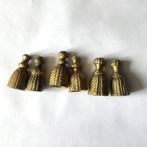 May include: A set of six gold-toned metal finials, each shaped like a decorative tassel with a textured, rope-like pattern on the body and a smooth, rounded top. These hardware pieces feature a hollow base and a metallic finish throughout.