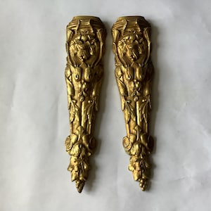 May include: A pair of matching gold-colored metal furniture appliqués featuring intricate relief carvings of floral and foliate motifs. Each elongated piece displays a symmetrical design with a tapered shape, detailed scrollwork, and leaf patterns.