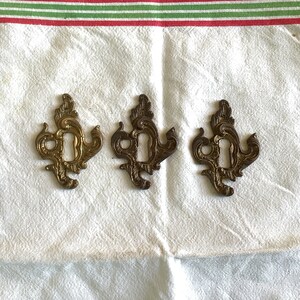 May include: Three ornate gold keyhole escutcheons with intricate designs. The escutcheons are arranged in a row on a white fabric background.