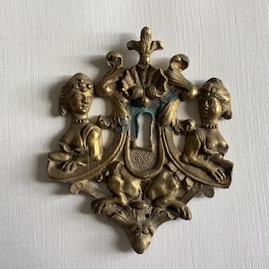 Könnte beinhalten: Antiker Messing-Schlüsselloch-Abdeckung mit aufwendigem Design. Es zeigt zwei gemeißelte weibliche Figuren, die ein Schlüsselloch flankieren. Das Messing hat eine goldene Farbe mit Spuren von Grünspan, die filigrane Details zeigen.