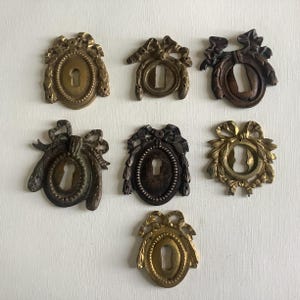 Könnte beinhalten: Sieben antike Schlüssellochabdeckungen aus Messing in verschiedenen Formen und Ausführungen. Die Abdeckungen weisen verzierte Designs mit Schleifen, Blättern und ovalen Rahmen auf. Die Schlüssellöcher sind oval geformt. Die Oberflächen reichen von Gold bis Dunkelbronze.